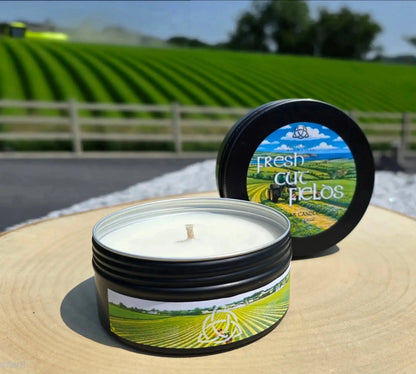 Fresh Cut Fields tin candle on a wood slice outdoors, green fields behind